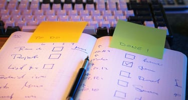 Handwritten to-do list in a notebook with a mechanical keyboard and colorful sticky notes.