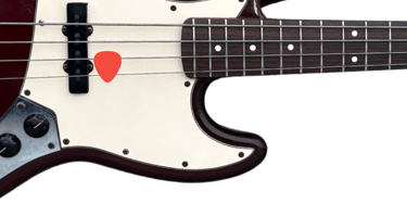 Close-up of a red electric bass guitar with a cream pickguard and an orange plectrum stuck in the strings.