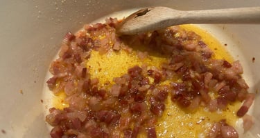 Diced pancetta browning in oil inside a white dutch oven with a wooden spoon stirring.