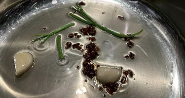 Sautéing garlic cloves, chili flakes, and scallion greens in oil inside a stainless steel pan.