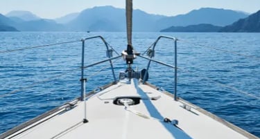 A white sailing yacht bow navigating blue ocean waters toward coastal mountains on a sunny day.