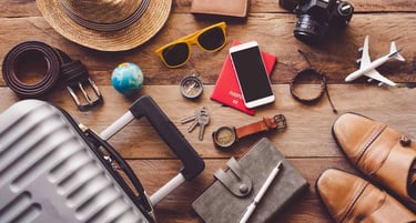 Travel essentials flat lay on a wooden table including a suitcase, passport, camera, and straw hat.