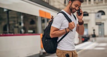 A smiling man with a backpack talking on a smartphone while waiting for a train.