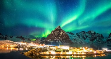 Vibrant green northern lights aurora borealis over a snowy mountain and glowing village in Lofoten, Norway.
