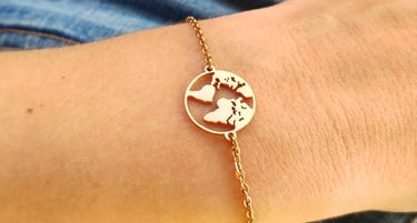 A gold world map charm bracelet worn on a wrist against a blue denim and yellow shirt background.