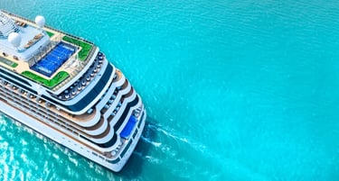 Aerial view of a luxury cruise ship sailing through turquoise ocean water with sports courts and sun decks visible.