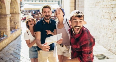 Four joyful friends on a sunny European city street, smiling and holding a map during their summer travel adventure.