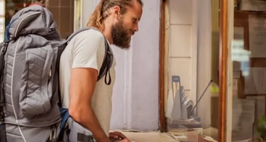 Male backpacker with a beard buying tickets at a train station counter during a solo travel trip.