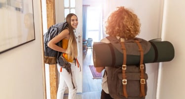 Two female backpackers with travel gear entering a bright hostel room for their budget adventure trip.