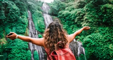 A female traveler with a red backpack overlooks a scenic tropical waterfall in a lush green jungle.