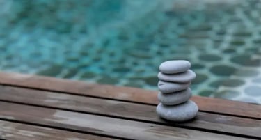 Balanced stack of smooth grey zen stones on a wooden deck by a tranquil blue swimming pool.