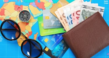 A brown leather wallet with Euro bills, credit cards, sunglasses, and a compass on a colorful world map.
