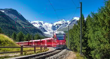 A red Bernina Express train traveling through the Swiss Alps with snow-capped mountains and green pine trees.