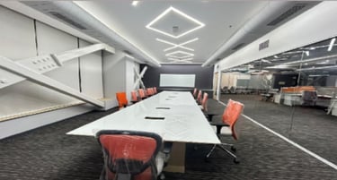 Modern conference room with a long white table, orange ergonomic chairs, and geometric LED ceiling lights.