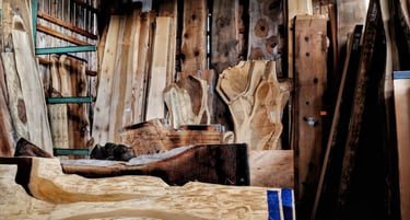 Assorted live edge wood slabs and reclaimed timber boards stacked in a woodworking lumber yard warehouse.