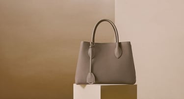 Elegante bolso de mano de piel color topo exhibido sobre un pedestal minimalista.