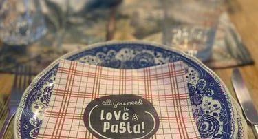 Rustic dinner table setting featuring blue patterned plates and a napkin reading All You Need Is Love and Pasta.