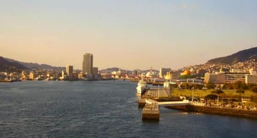Navio de cruzeiro chegando ao porto de Nagasaki, com pessoas aguardando na orla e colinas verdes ao fundo