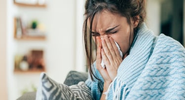 A woman sneezing into a tissue while wrapped in a blue blanket, suffering from cold or allergy symptoms.