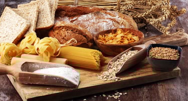 An assortment of whole grain foods including rustic bread, pasta, oats, and wheat stalks on a wooden board.