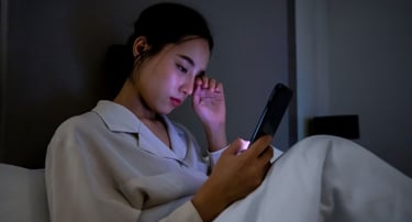 A young woman in bed rubbing her tired eyes while looking at a smartphone screen in a dark room.