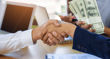Business partners shaking hands over a contract while a woman holds stacks of US dollar bills.