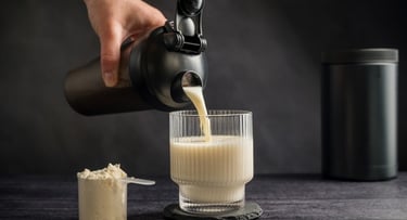 Hand pouring a vanilla protein shake from a black shaker bottle into a ribbed glass with supplement powder.