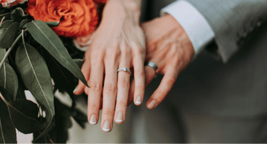 Close-up of a married couple's hands displaying a diamond engagement ring and wedding bands next to a bridal bouquet.