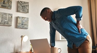 A man standing at a desk holding his lower back in pain while working on a laptop at home.
