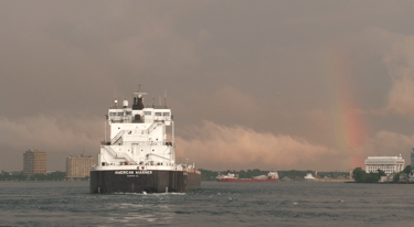 Stern of a black and white freighter as it sails towards a rainbow.