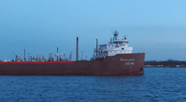 Front section of a classic forward pilothouse red and white freighter passing an oil refinery.