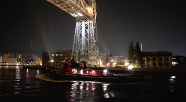 A Classic red and green tugboat in the dark, passing under the lit-up Duluth Aerial Lift Bridge.