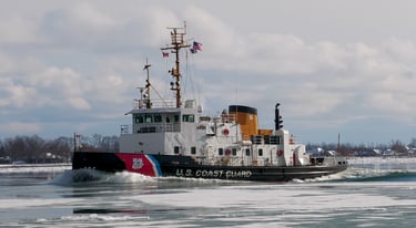 A Black and white U.S. Coast Guard tugboat making a large wake on icy water.