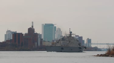 Right-side stern view of a gray navy ship passing some high-rise buildings.