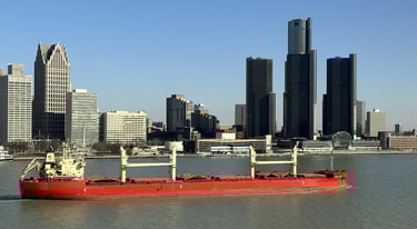 A red oceangoing ship with tan deck cranes passing downtown Detroit.