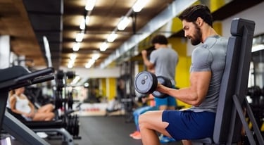 A muscular man performing seated bicep curls with dumbbells at a modern gym.