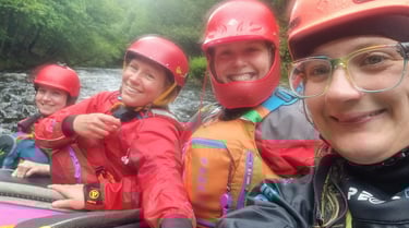 4 women perched on a rock in the middle of the river in white water kayaks. 