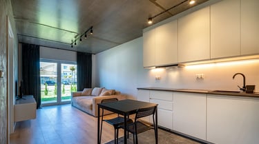 Nowoczesny salon z aneksem kuchennym: białe szafki, stół z krzesłami, sofa, drzwi balkonowe.