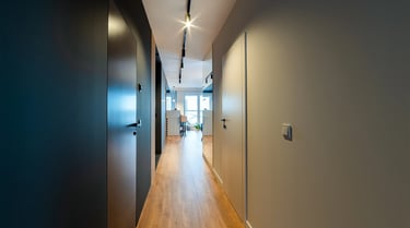 Korytarz z drzwiami po obu stronach, drewniana podłoga, listwowe oświetlenie sufitowe okno na końcu.