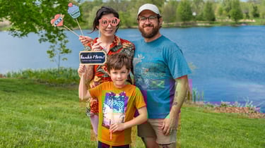 Staged photo of a couple with a young child in front of a lake, holding up paper props