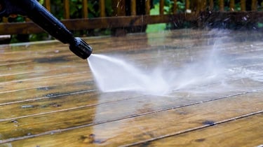 A high-pressure washer nozzle spraying water to clean a dirty wooden deck outdoors.