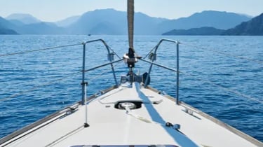 View from the bow of a white sailboat cruising on blue ocean water toward distant mountains.