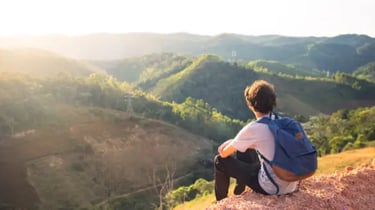A male hiker with a blue backpack sits on a hill at sunset, looking over a lush mountain valley.