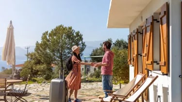 A smiling female traveler with a suitcase greeting a host outside a Mediterranean vacation rental.