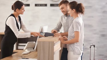 A couple uses a credit card to check in at a hotel reception desk with a smiling concierge.