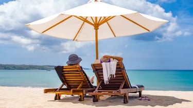 Two people relaxing on wooden lounge chairs under a white beach umbrella by the turquoise ocean.