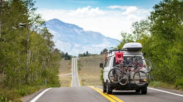 White camper van with mountain bikes driving on a scenic road toward distant mountains.