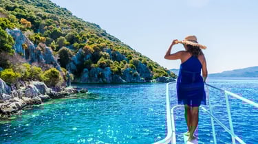 Woman in a blue dress and straw hat on a boat deck overlooking a tropical coastline with turquoise water.
