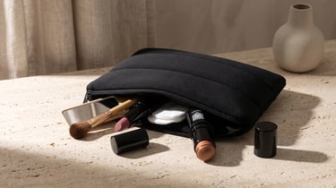 Black quilted makeup bag with lipstick, brush, and cosmetics on a stone surface.
