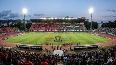 Estádio do La Guaira lotado para jogo contra o Fluminense pela Libertadores, com campo iluminado e torcida vibrante.